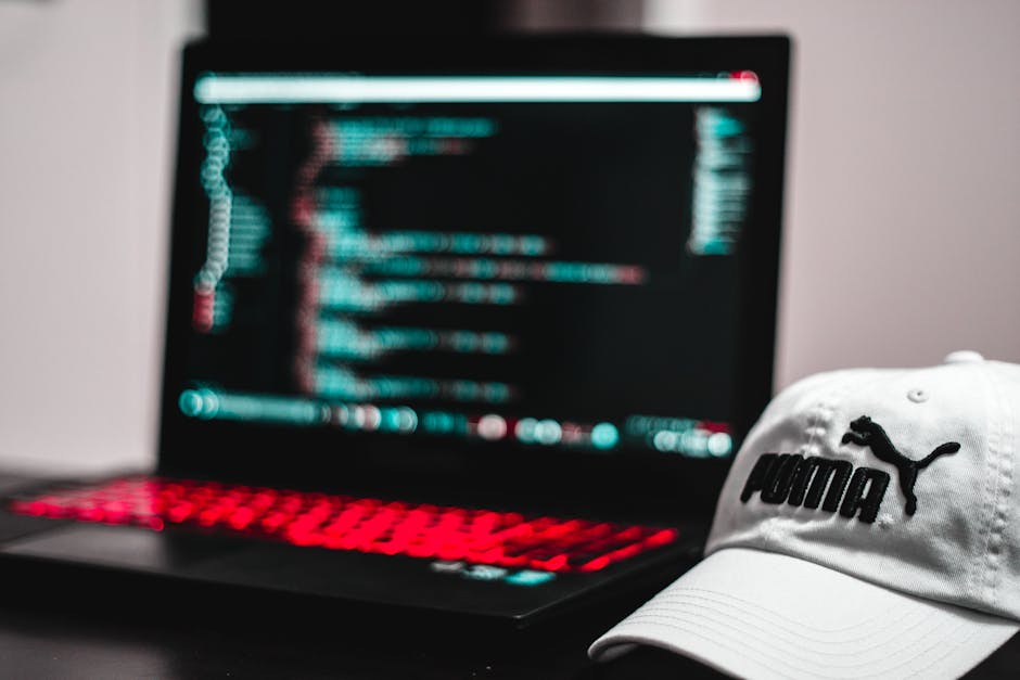 The image features a black laptop with a glowing red keyboard, displaying lines of code or text on its screen, which is slightly blurred to emphasize the digital content. In front of the laptop is a w