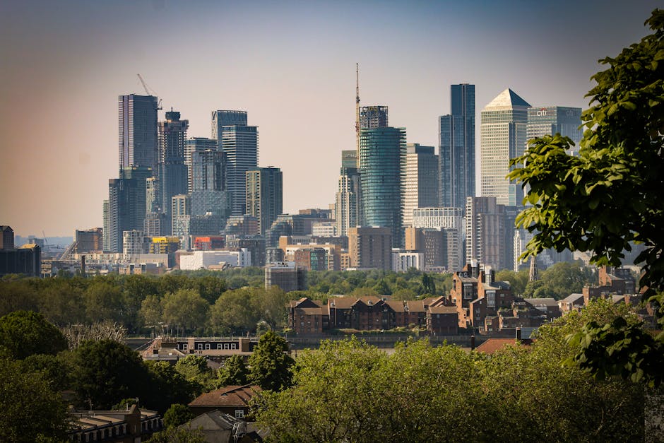 A panoramic view of London's city skyline featuring tall modern skyscrapers with glass facades reflecting sunlight, including iconic high-rise buildings under construction, set against a clear sky. In