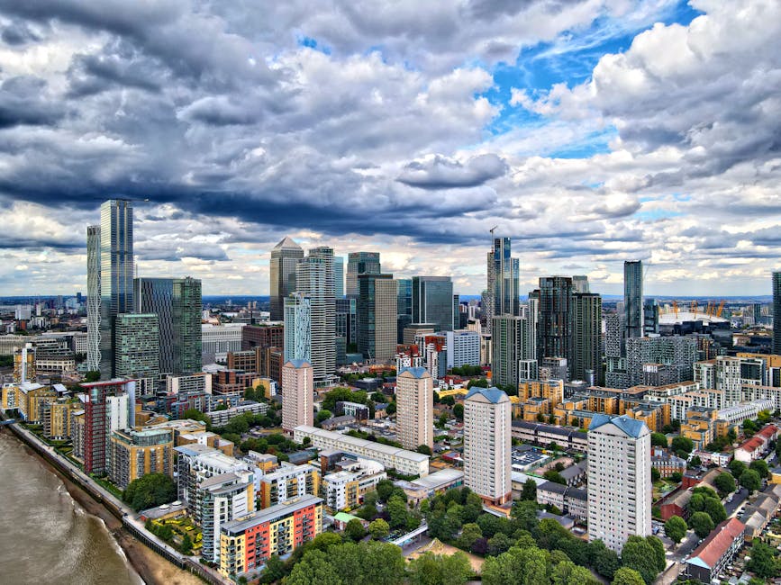 A panoramic view of London's city skyline featuring tall modern skyscrapers with glass facades reflecting sunlight, including iconic high-rise buildings under construction, set against a clear sky. In